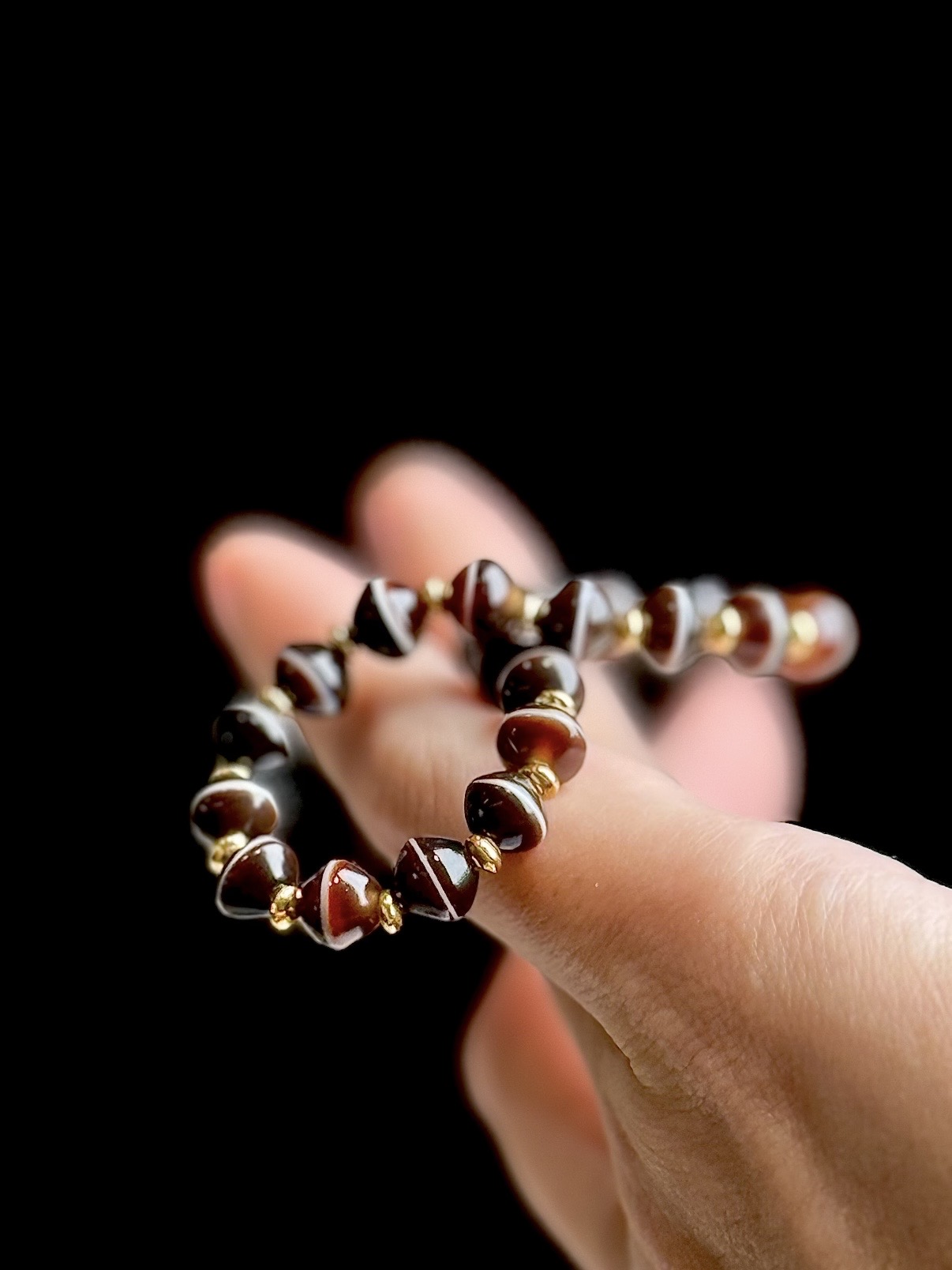 A banded agate bracelet of dark red bicone beads with white bands and gold rondelles, held over the fingers of a hand against black.